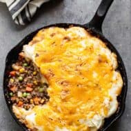Overhead photo of a cast iron pan with baked cottage pie consisting of a meat and vegetable gravy mixture topped with mashed potatoes and melted cheddar cheese.