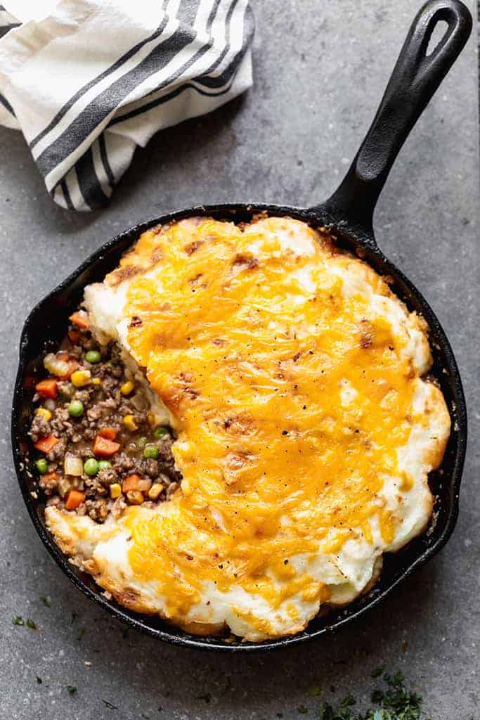 Overhead photo of a cast iron pan with baked cottage pie consisting of a meat and vegetable gravy mixture topped with mashed potatoes and melted cheddar cheese.