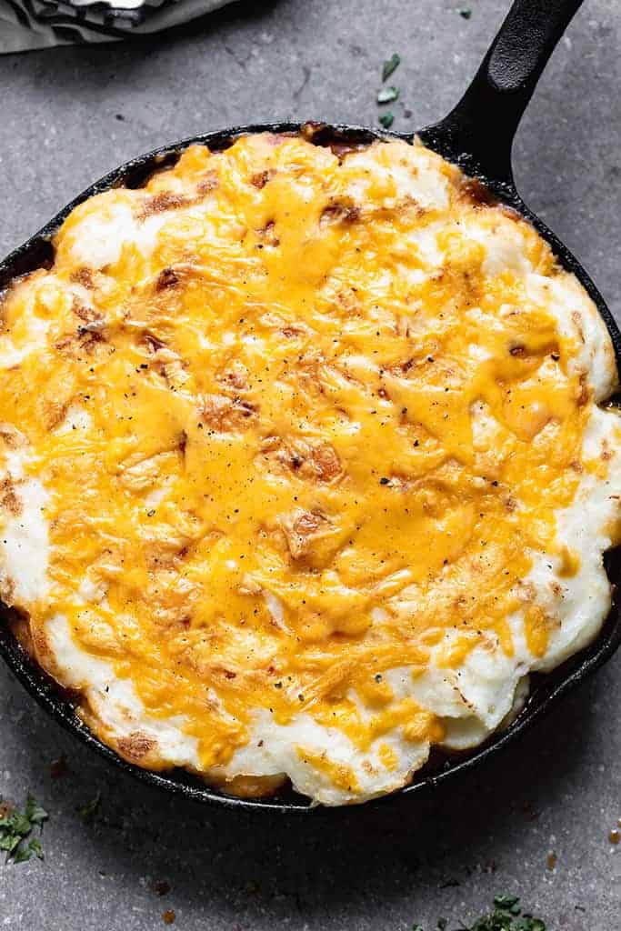 Baked cottage pie with cheddar cheese melted on top of a layer of mashed potatoes inside a cast iron pan.