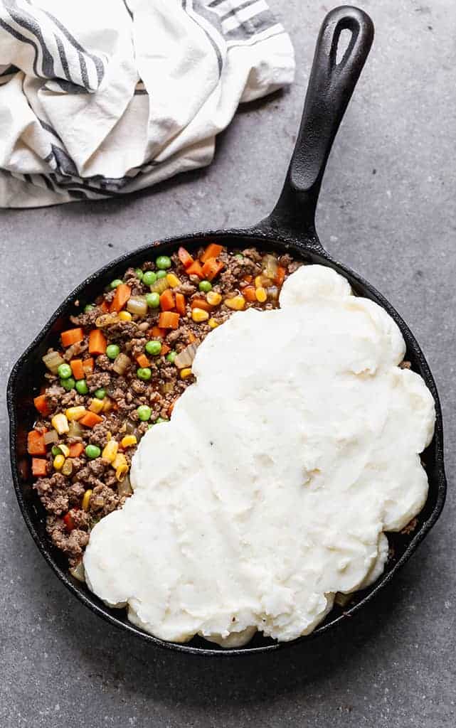 A cast iron skillet with the filling for cottage pie and half of the pan with mashed potatoes smoothed on top.