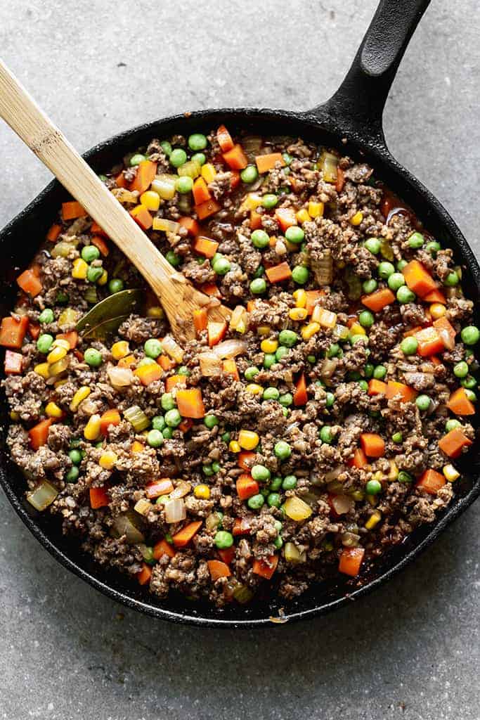 A cast iron skillet with a mixture of cooked ground beef, carrots, peas, corn and a gravy sauce.