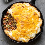 An easy Cottage Pie recipe in a cast iron pan with some meat and vegetable gravy showing under the layer of cheesy mashed potatoes.