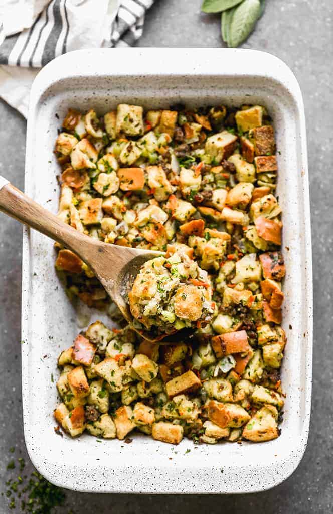 Sausage stuffing in a baking dish with a wooden spoon lifting up a serving.