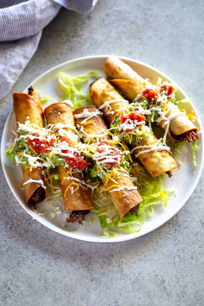 Five beef taquitos fanned out on a plate topped with guacamole, sour cream and salsa.