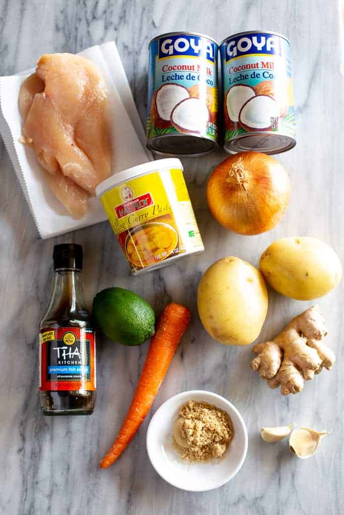 A marble board with the ingredients for yellow curry including curry paste, coconut milk, chicken, potatoes. onion, carrot, lime, ginger, garlic, fish sauce and brown sugar.