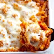 A white casserole dish with ziti pasta, meatballs and cheese lifted out with a spoon.