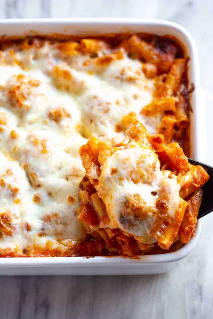 A white casserole dish with ziti pasta, meatballs and cheese lifted out with a spoon.