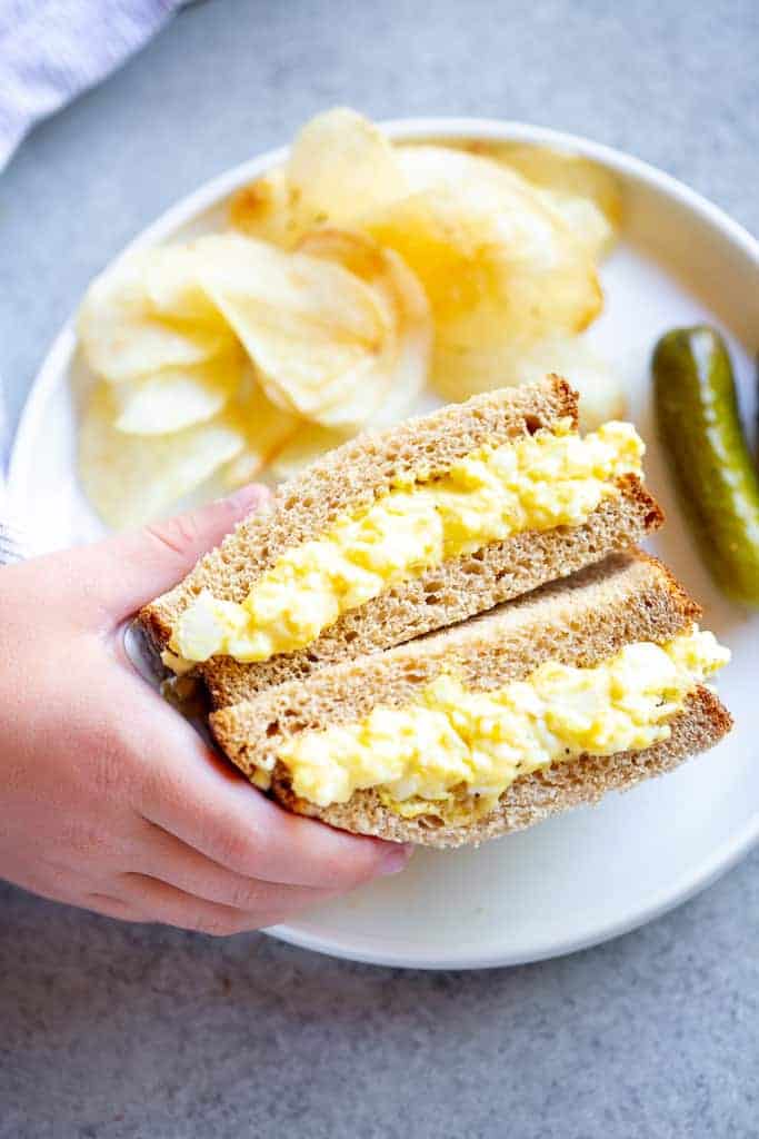 An egg salad sandwich on a plate served with pickles and potato chips.