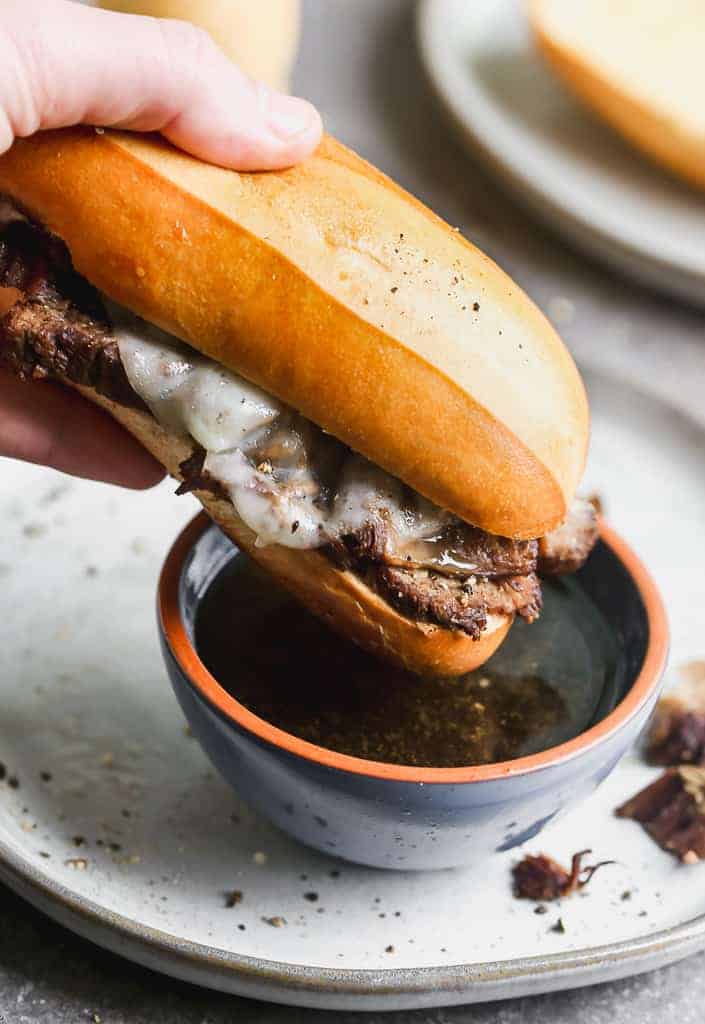 A french dip sandwich being dipped into a small bowl of au jus. 