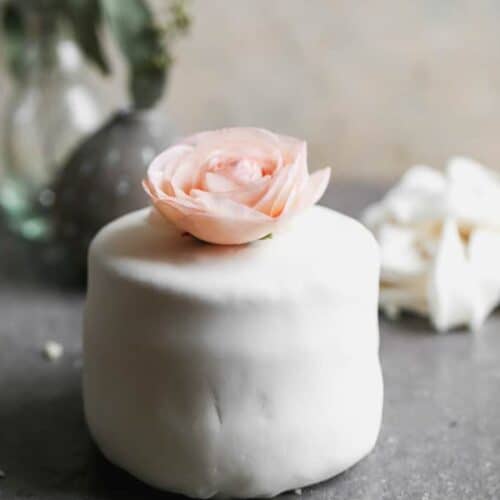 A small cake covered with white marshmallow fondant and a flower on top.