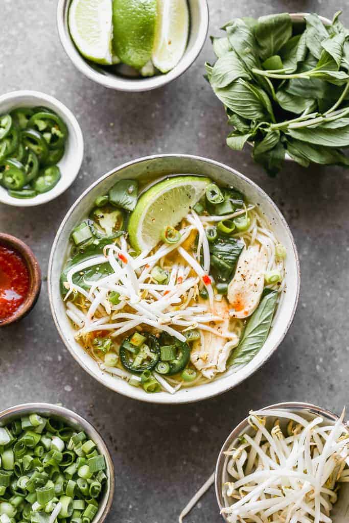 A bowl of Pho soup with chicken, cilantro, onion, basil, bean sprouts and a lime wedge.