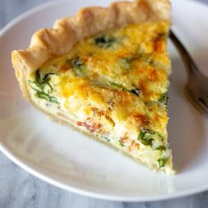 A slice of spinach and bacon quiche on a plate with a fork.