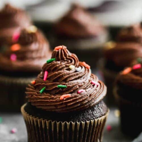 Chocolate cupcakes with buttercream frosting and sprinkles on top, lined up on a grey board .