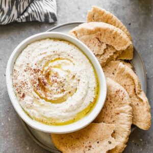 A bowl of hummus set on a plate with pita bread around it.