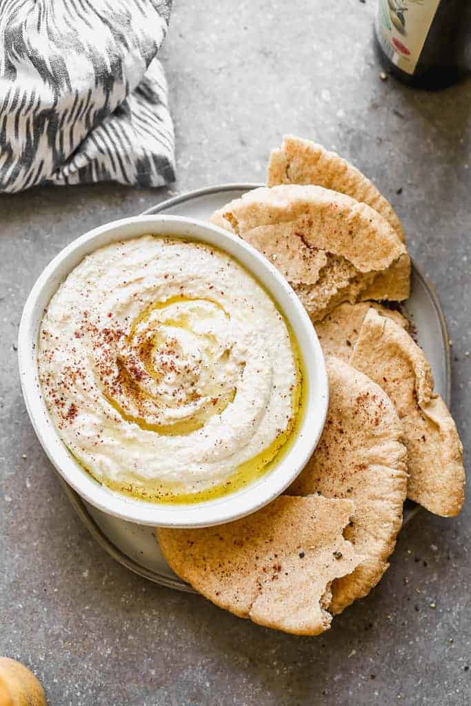 A bowl of hummus set on a plate with pita bread around it. 
