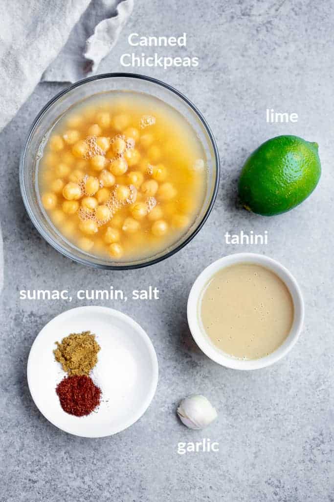 The ingredients for hummus, labeled, including canned chickpeas, lime, tahini, garlic, sumac, salt, and cumin.