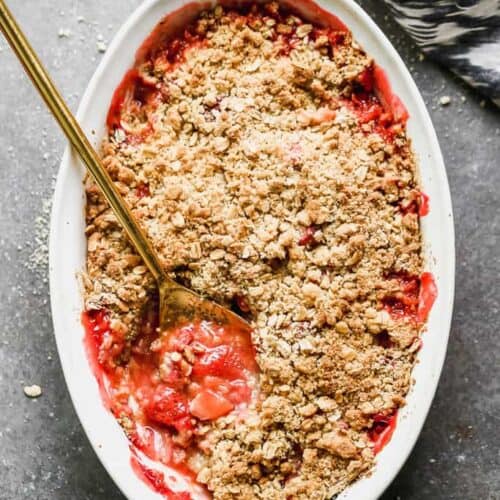 An oval baking dish with strawberry rhubarb crisp and a spoon in it for serving.