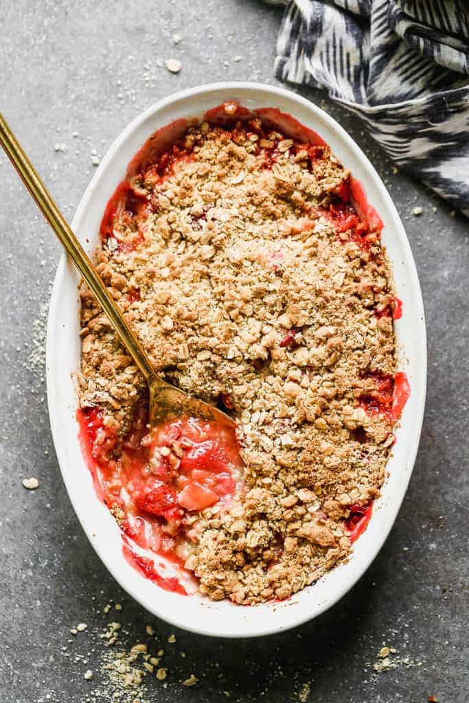 An oval baking dish with strawberry rhubarb crisp and a spoon in it for serving.