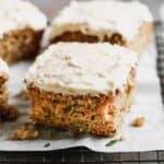 Slices of zucchini cake with cream cheese frosting on parchment on a wire rack.