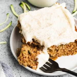 A piece of the best Zucchini Cake recipe served on a white plate with a cream cheese frosting spread on top and a bite on the fork.