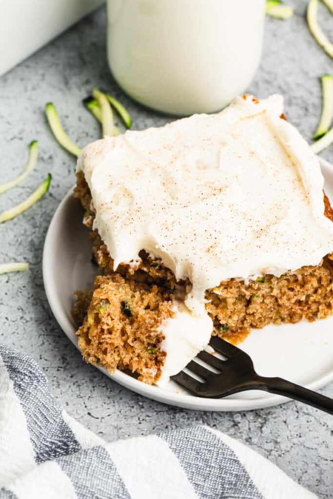 A piece of the best Zucchini Cake recipe served on a white plate with a cream cheese frosting spread on top and a bite on the fork.