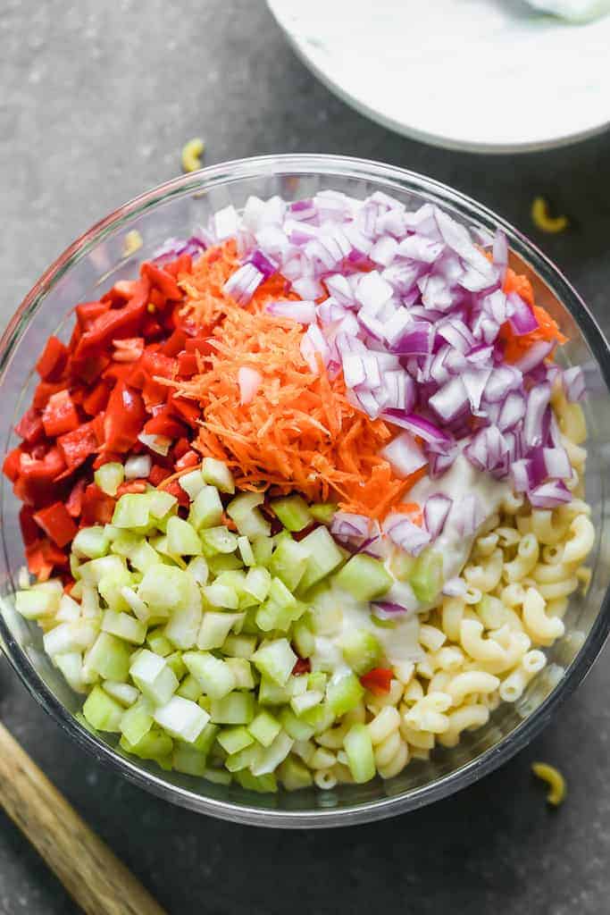 Des nouilles coudées cuites et des légumes hachés s'ajoutent dans un bol pour faire une salade de macaroni.