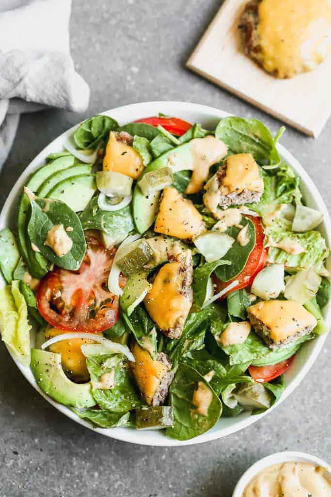 Cheeseburger salad in a white bowl, topped with tomato, avocado, onion and dressing.