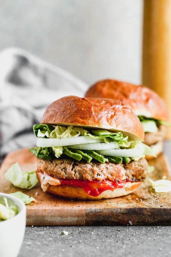 Two turkey burgers served in buns, topped with avocado, lettuce and onion.