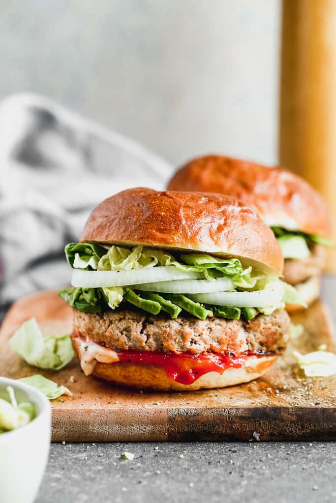 Two turkey burgers served in buns, topped with avocado, lettuce and onion.