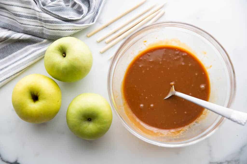 Melted caramel in a bowl with apples and dipping sticks on the side to make caramel apples.