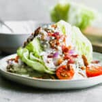 An easy wedge salad on a plate with blue cheese dressing, bacon, onion, and tomatoes.