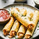 Five chicken taquitos lined on a plate with a bowl of salsa on the side.