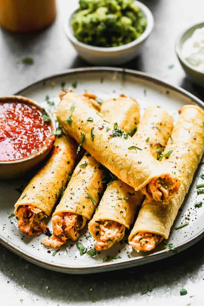 Five chicken taquitos lined on a plate with a bowl of salsa on the side.
