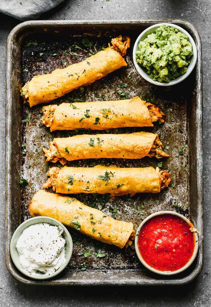 Chicken taquitos on a tray with bowls of guacamole, salsa and sour cream.