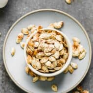 A bowl of roasted pumpkin seeds sitting on a plate, with some seeds spilling over onto the plate.