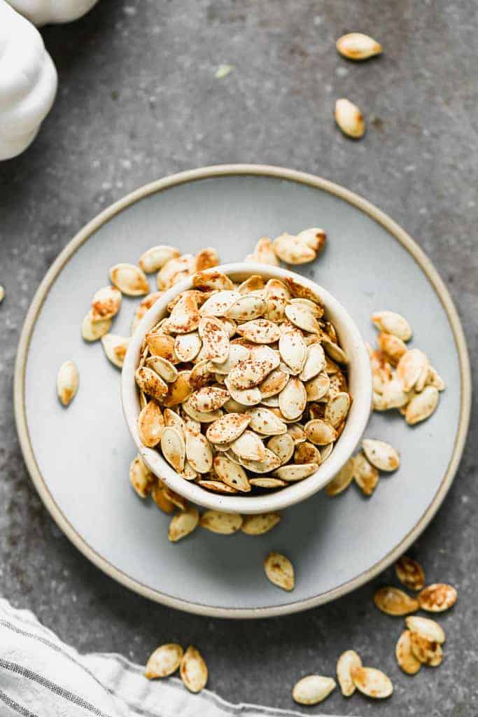 A bowl of roasted pumpkin seeds sitting on a plate, with some seeds spilling over onto the plate.