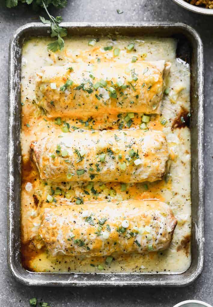 Three burritos on a baking tray smothered in green chile sauce and topped with cheese and green onion.