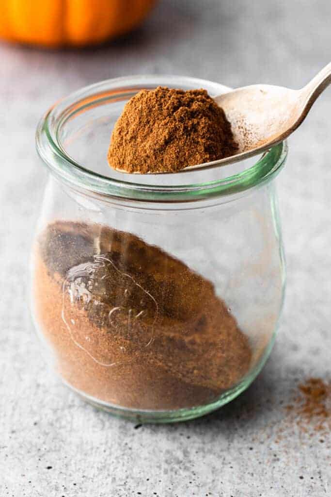 Homemade pumpkin pie spice in a clear glass jar with a spoon lifting some out.