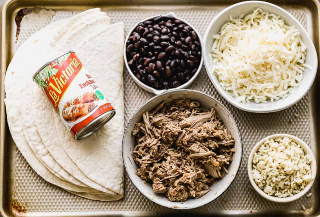 The ingredients needed to make wet burritos, including flour tortillas, enchilada sauce, shredded pork, black beans, cooked rice and shredded cheese.
