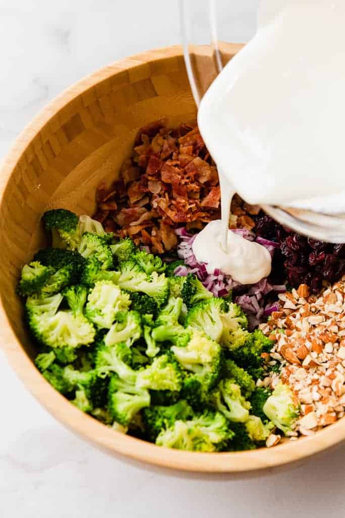 Broccoli salad ingredients in a bowl and dressing being poured on top.