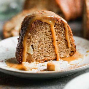 A slice of a fresh Apple Bundt Cake recipe served on a white plate and topped with a homemade caramel sauce.