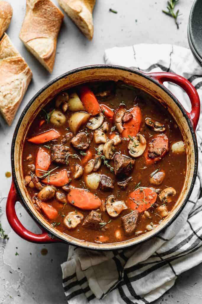 A large pot of beef stew with carrots, potatoes and mushrooms and sliced baguette on the side.