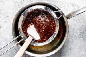 Mélange de sauce chili rouge poussé à travers une passoire à mailles fines dans une casserole.