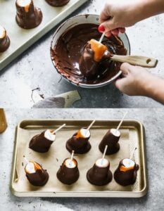 Large marshmallows on a stick being dipped in melted chocolate, then place on a lined sheet.