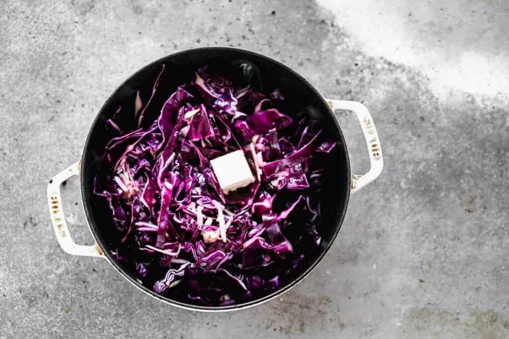 A white dutch oven pot with shredded purple cabbage, grated apple and butter on top, ready to cook.