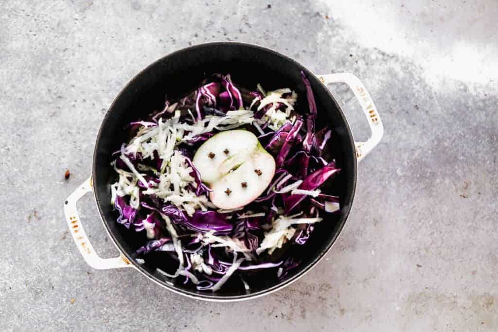 Shredded red cabbage and apple, and a half of an apple with whole cloves in it, in a large pot.