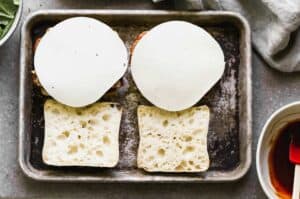 Ciabatta bread rolls cut in half on a baking tray, topped with a slice of meatloaf and provolone cheese.