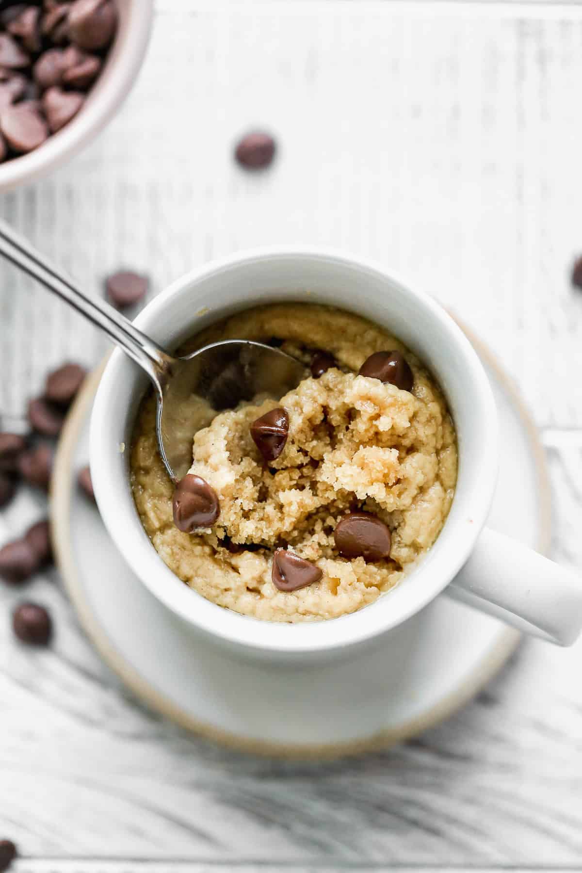 The best Peanut Butter Mug Cake recipe in a white mug with a few chocolate chips on top and a spoon scooping up a bite.