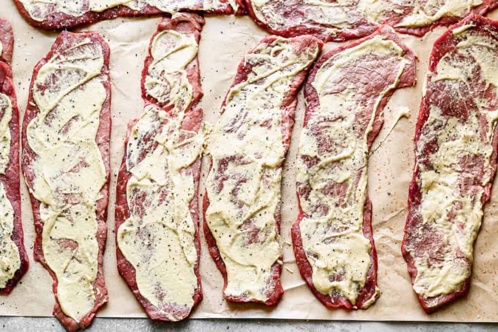 Thin sliced of round roast laid on parchment paper with mustard smoothed on top, to make Rouladen.