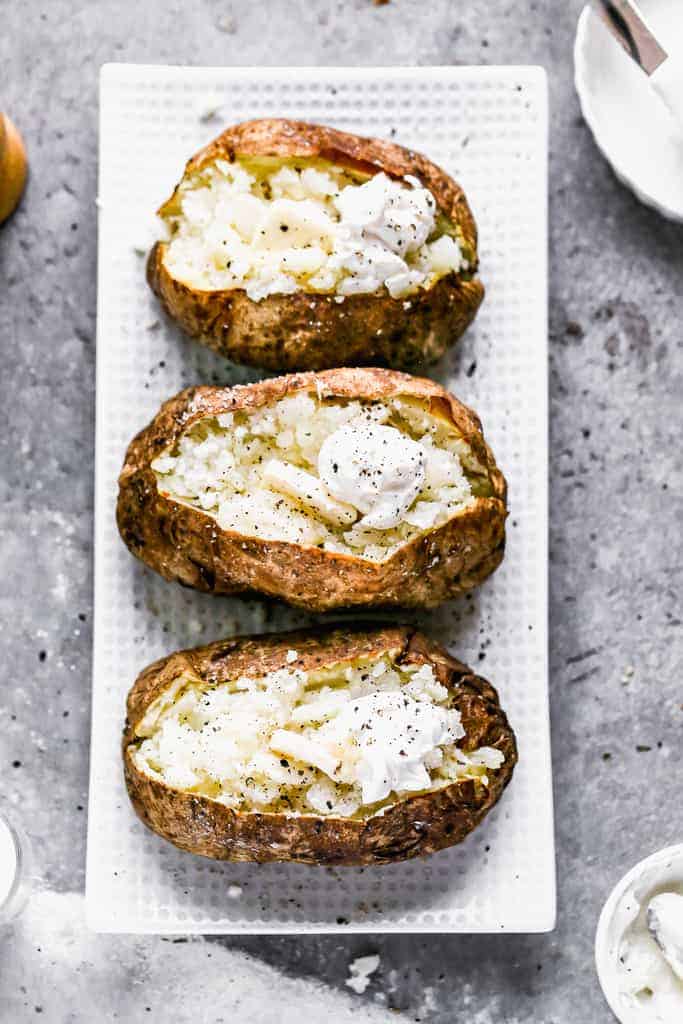 Three Air Fryer Baked Potatoes on a serving dish with the tops of the potatoes slit open and butter and sour cream added to the center.